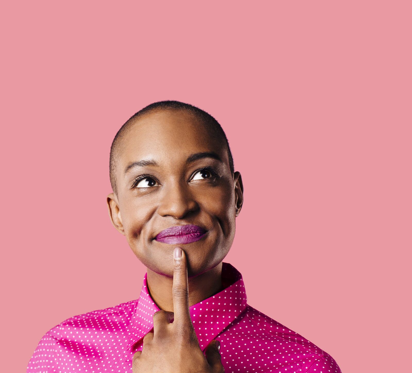 Portrait of a young woman in pink shirt with finger on mouth looking up thinking, isolated on pink studio background