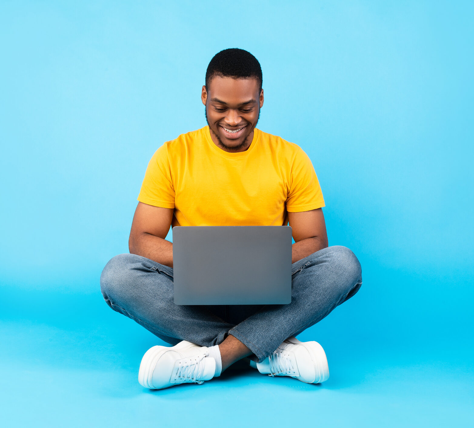 Young man in yellow shirt sitting cross legged happily looking down at laptop with blue background.