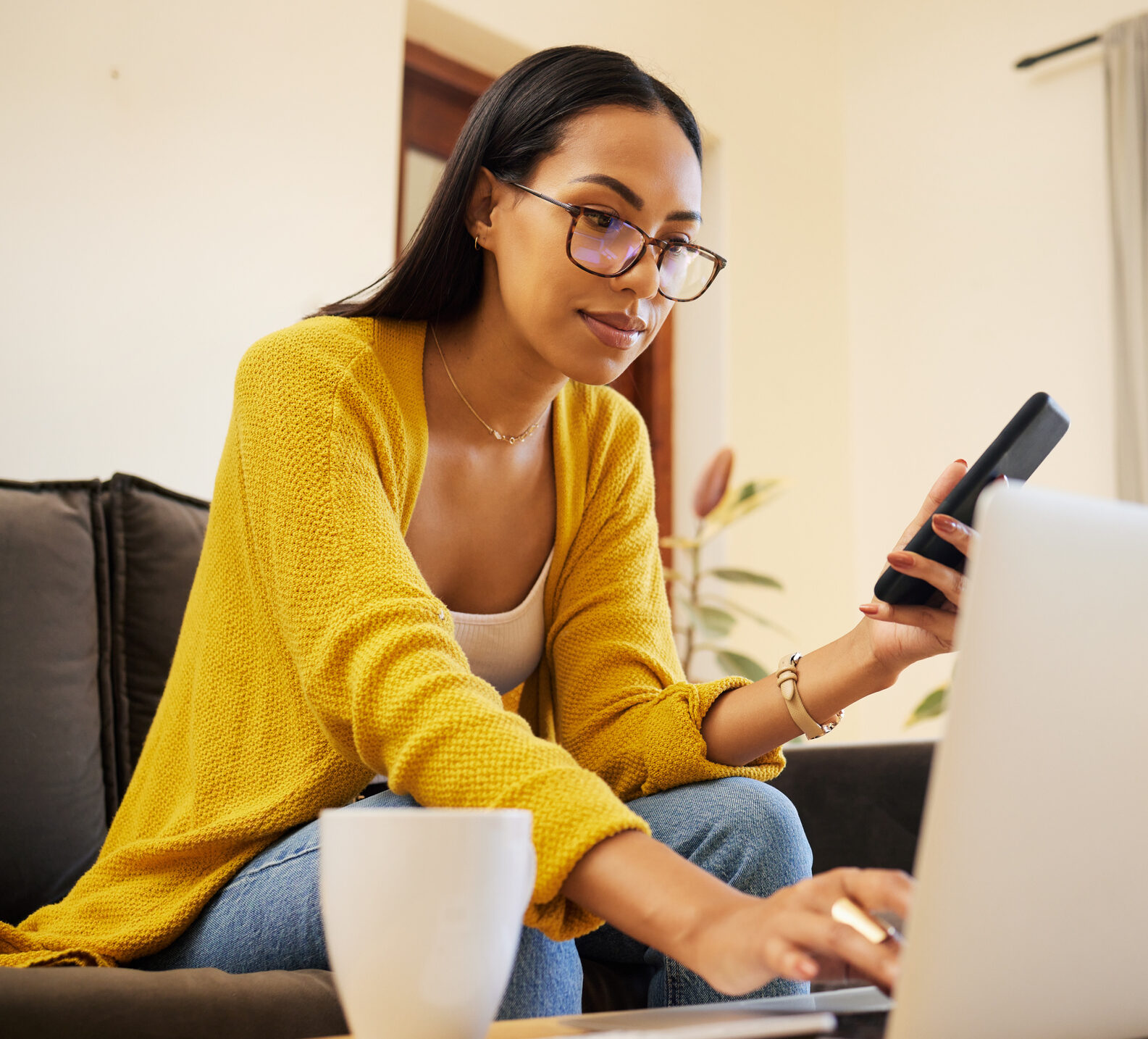 Woman working remote while typing on her laptop and holding her smartphone sitting on a sofa in a bright living room. One focused young hispanic female with glasses at home using modern technology Time in range insurance coverage