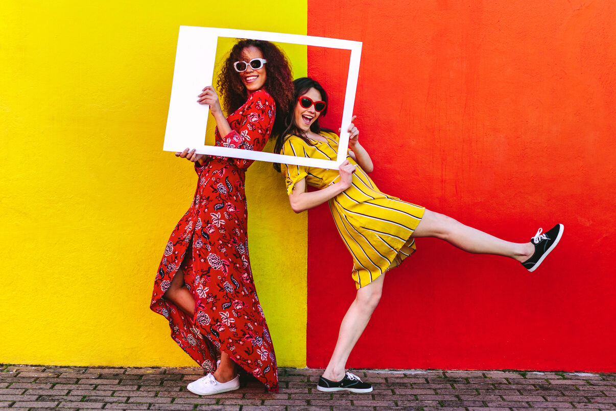 Tow women with sunglasses having fun with a giant picture frame against a colorful wall