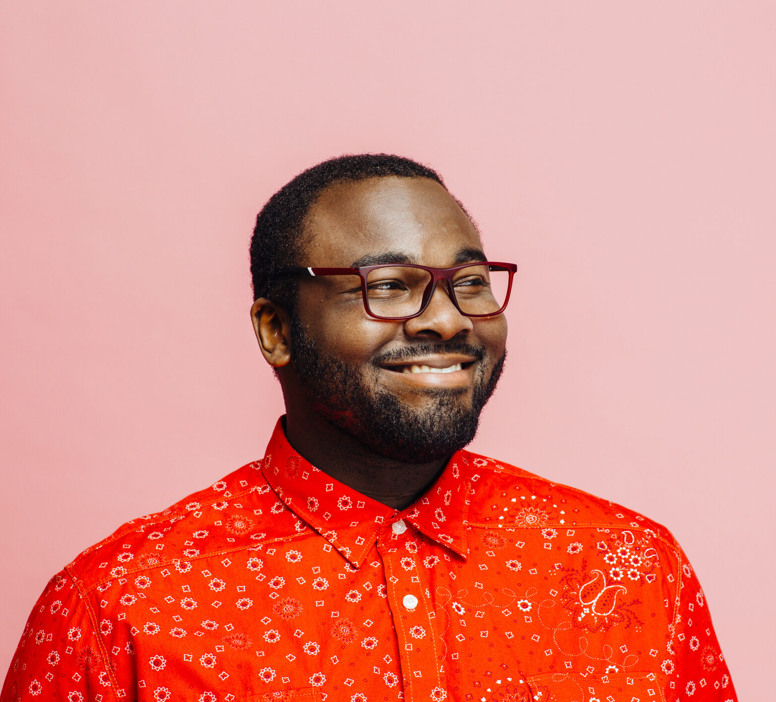 Man in bright red shirt and glasses smiling and looking off camera against pink background. He is happy to learn about time in range