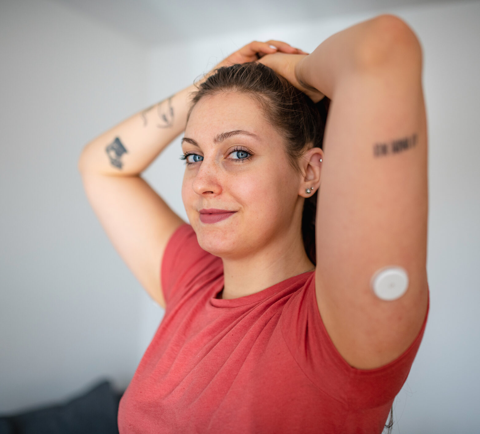 Partial side view of Caucasian woman in casual clothing with glucose sensor on her arm and hands on her head smiling at camera.